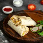 side view of shaurma kebab on a wooden board with sauces on black background